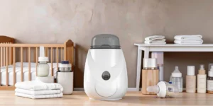 A compact white electric baby bottle warmer and steam sterilizer sitting on a kitchen counter, with a clear baby bottle placed inside the unit ready for warming.